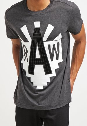 Osoba nosząca ciemnoszary t-shirt z dużym czarno-białym nadrukiem przedstawiającym litery R, A i W na przodzie.