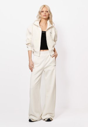 Egy szőke nő áll, krémszínű cipzáras dzsekit, hozzá illő bőszárú nadrágot, fekete crop topot és sportcipőt viselve, egyszínű fehér háttér előtt.