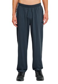 Donkere grijze sportbroek van een geweven stof, met een elastische tailleband en een zijzak. Zwart logo aan de linkerkant. G gedragen met zwarte schoenen.