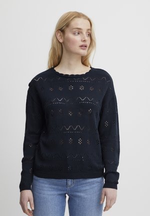 Maglione lavorato a maglia blu navy con motivi a trafori, scollatura ondulata e polsini a coste, abbinato a jeans azzurri chiari.