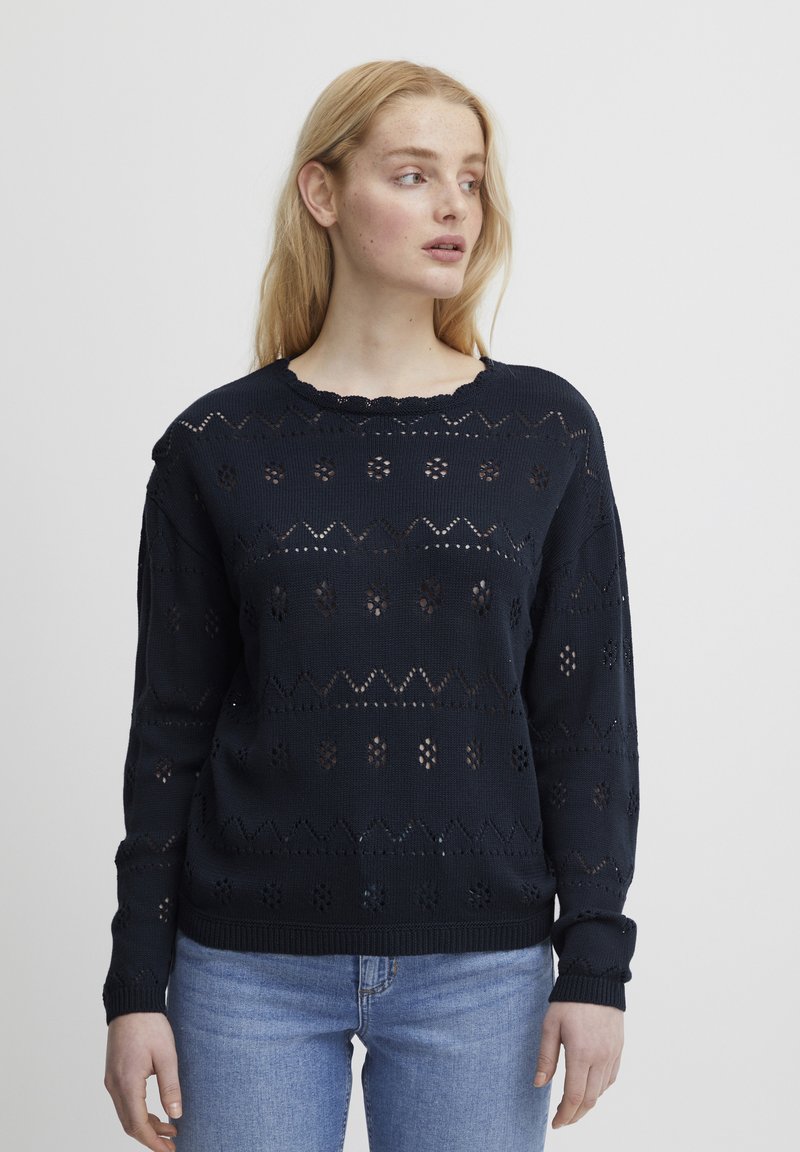 Granatowy sweter dzianinowy z ażurowymi wzorami, falbaniastym dekoltem i ściągaczami, w połączeniu z jasnoniebieskimi dżinsami.