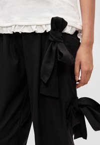 Calças de tecido preto com grandes laços nas laterais, combinadas com uma blusa branca com barra franzida. A vista em close destaca as texturas.