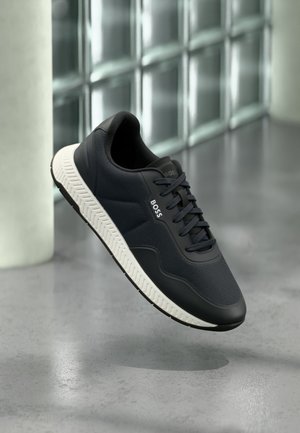 Baskets noires BOSS avec semelle blanche, dessus en mesh et cuir, flottant au-dessus d'un sol en béton poli près d'un mur en blocs de verre.