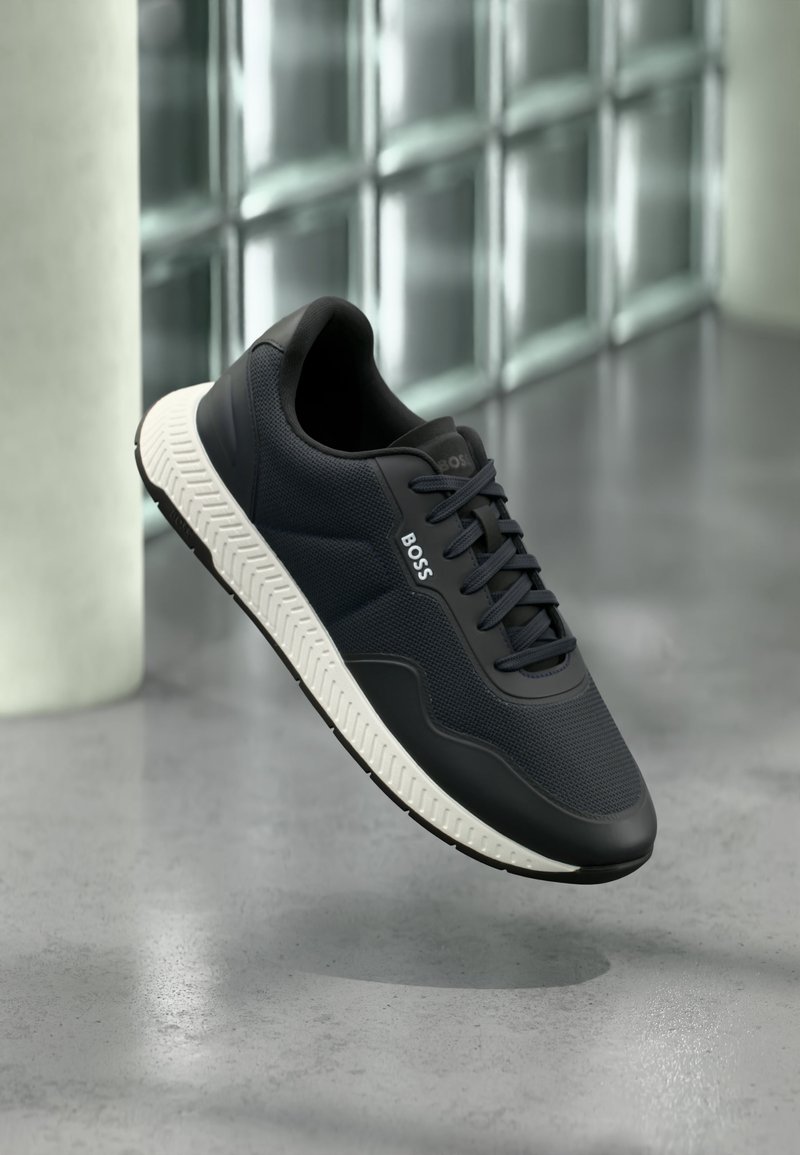 Baskets noires BOSS avec semelle blanche, dessus en mesh et cuir, flottant au-dessus d'un sol en béton poli près d'un mur en blocs de verre.