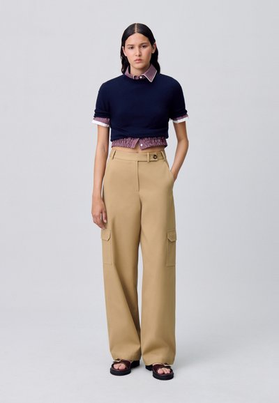 Maglione cropped blu navy sopra una camicia a scacchi rossa, abbinato a pantaloni cargo beige a gamba larga con tasche laterali e chiusura con bottone. Sandali neri.