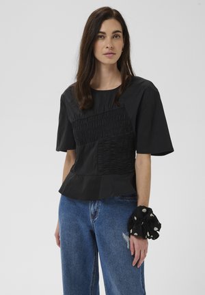 Vrouw met lang donker haar die een zwarte textuur korte mouw top, blauwe spijkerbroek en een zwarte scrunchie met witte stippen om haar pols draagt.