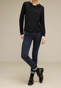 Kvinna som bär en svart långärmad blus, mörkblå uppviktajeans, svarta strumpor och svarta snörskor, stående mot en enfärgad bakgrund.