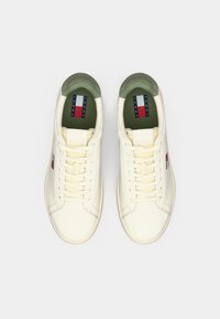 Cream-colored sneakers with a smooth leather finish, featuring a green padded collar and a logo tag on the tongue. Classic lace-up design.