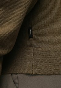 Sötét olíva zöld kötött pulóver bordázott textúrával, amelyen a varrásnál egy kis fekete címke található "PREMIUM" felirattal.