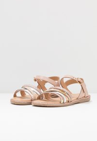 Sandalias de puntera abierta con un upper de cuero sintético rosa pálido, que presentan tiras brillantes en dorado y plateado, y una hebilla ajustable en el tobillo. Suela plana.