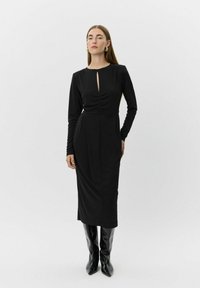 Kvinde iført en sort langærmet midi-kjole med en frontslids, stylet med sorte knæhøje støvler og guld statement-øreringe.