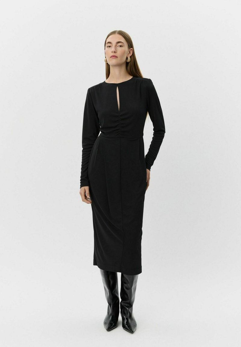 Kvinde iført en sort langærmet midi-kjole med en frontslids, stylet med sorte knæhøje støvler og guld statement-øreringe.