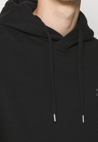 Černá mikina s kapucí, se šňůrkou s kovovými konci, měkkou texturou látky a malým vyšitým logem na hrudi.