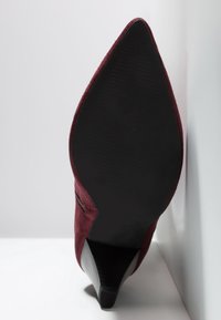 Zapato de tacón alto de ante burdeos con punta fina, suela texturizada negra y tacón apilado, fotografiado desde el lado de la suela sobre un fondo blanco.