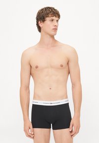 Schwarze Baumwoll-Boxershorts mit einem weißen Taillenbund, der das Logo "TOMMY HILFIGER" trägt. Der Stoff wirkt glatt und eng anliegend.