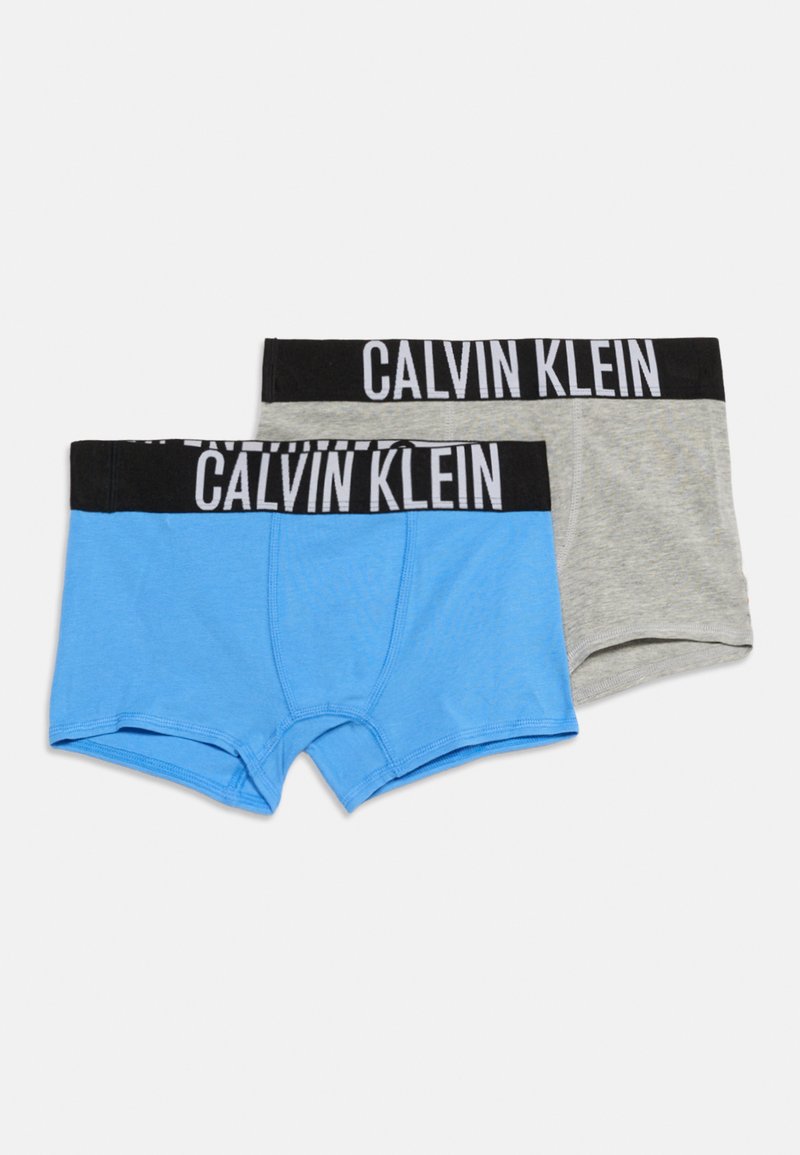 Δύο ζευγάρια ανδρικά μπόξερ Calvin Klein για αγόρια, ένα ανοιχτό μπλε και ένα ανοιχτό γκρι, και τα δύο με μαύρες ζώνες μέσης και λευκά λογότυπα της εταιρείας.