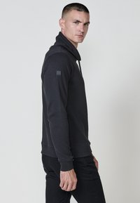 Felpa nera con texture, colletto alto e zip laterale. Le maniche lunghe e i polsini a costine aumentano il comfort. Piccola patch logo sulla spalla.