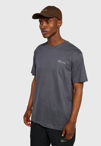 Szara bawełniana koszulka z krótkimi rękawami, okrągłym dekoltem i nadrukiem na lewej piersi. Połączona z czarnymi szortami i brązową czapką.