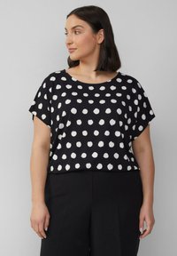 Vrouw met schouderlang, donker haar, die een zwart shirt met witte stippen en een zwarte broek draagt, terwijl ze naar links kijkt.
