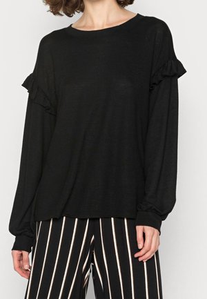 Kobieta ubrana w czarną bluzkę z długim rękawem z falbanami na rękawach oraz czarne spodnie o szerokich nogawkach w białe pionowe paski.