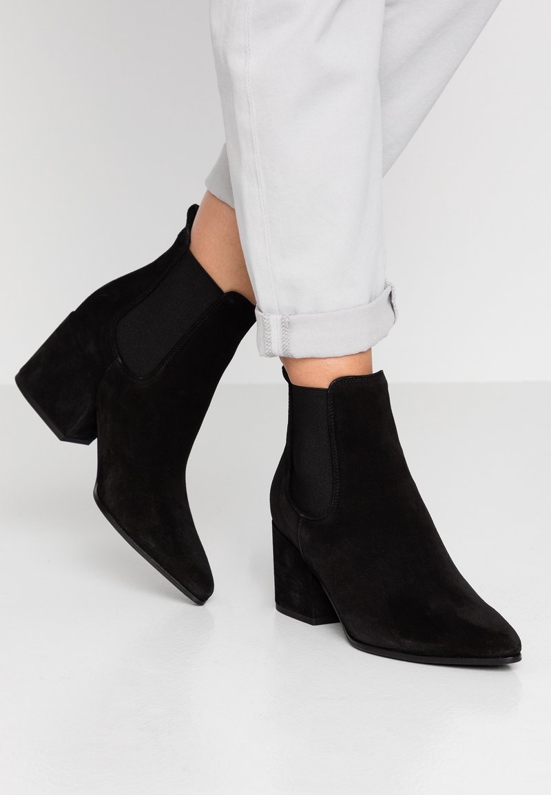 Bottines en daim noir avec talons carrés portées avec un pantalon gris clair remonté sur un sol et un fond blancs.