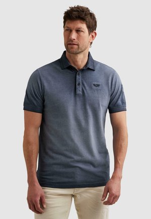 Mann mit kurzen braunen Haaren, der ein blaues Polo-Shirt mit kurzen Ärmeln und beige Hose trägt, steht vor einem schlichten hellen Hintergrund.
