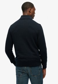 Homme vu de dos portant un pull bleu marine foncé avec un col replié et un jean denim bleu sur un fond blanc.