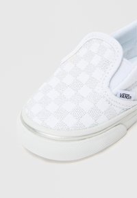 Fehér slip-on sportcipő, texturált sakkmintával, gumitalppal és egy kis márkajelzéssel az oldalán. Könnyű anyagból készült felsőrész.