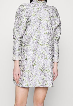 Femme portant une robe courte à col montant avec des manches bouffantes, un motif floral en lavande et vert, et des poignets ornés de perles.