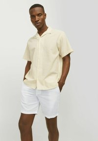 Camisa de botones amarilla claro de manga corta con sutiles rayas verticales, combinada con pantalones cortos blancos. Textura suave, corte relajado.