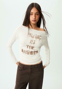 Krémový top s dlouhým rukávem a odkryté ramena s potiskem "MUSIC IS THE ANSWER" v hnědé barvě. Spárovaný s tmavě hnědými upnutými kalhotami.