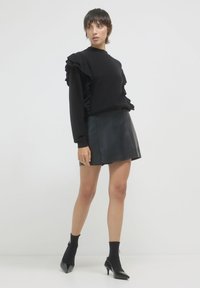 Pull noir avec des manches volantes, associé à une mini-jupe en cuir noir et des bottines à chaussettes noires. Textures lisses et silhouette ajustée.