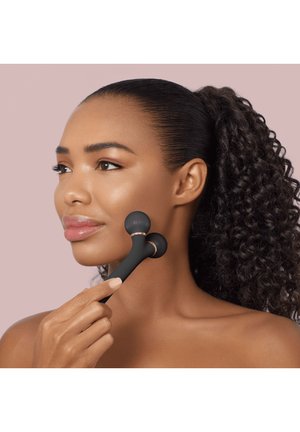 Woman with curly hair uses black dual-ball facial massage tool on her cheek against a light pink background.