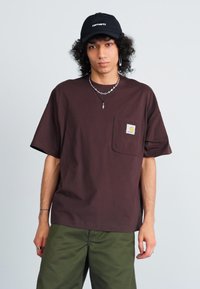 Brūna kokvilnas T-kreklu ar kabatu, īsām piedurknēm un Carhartt birku. Saskaņots ar olīvzaļām biksēm un papildināts ar melnu cepuri.