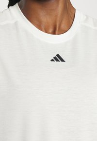 Biela bavlnená tričko s okrúhlym výstrihom, so čiernym logom Adidas v strede. Tkanina má hladkú textúru a jemné variácie.