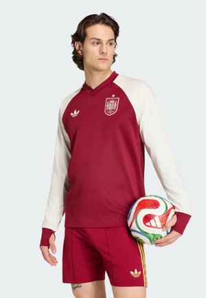 Ung mand iført rød og hvid spansk fodboldtrøje og shorts holder en farverig Adidas fodbold og kigger til siden mod en ensfarvet baggrund.