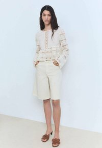Blusa color crema con pizzo, maniche lunghe, abbinata a pantaloni corti beige chiaro. Sandali con tacco marroni con dettagli floreali completano l'outfit.