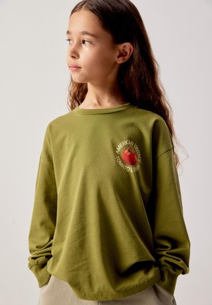 Meitene ar gariem brūniem matiem, valkājot olīveļļas zaļu sporta kreklu ar sarkanu tomātu un tekstu "American Vintage Tomato Lover", skatās pa kreisi.