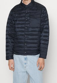 Mężczyzna w ciemnoniebieskiej pikowanej kurtce zapinanej na guziki z kieszenią na piersi oraz jasnoniebieskich dżinsach, stojący na jednolitym tle.