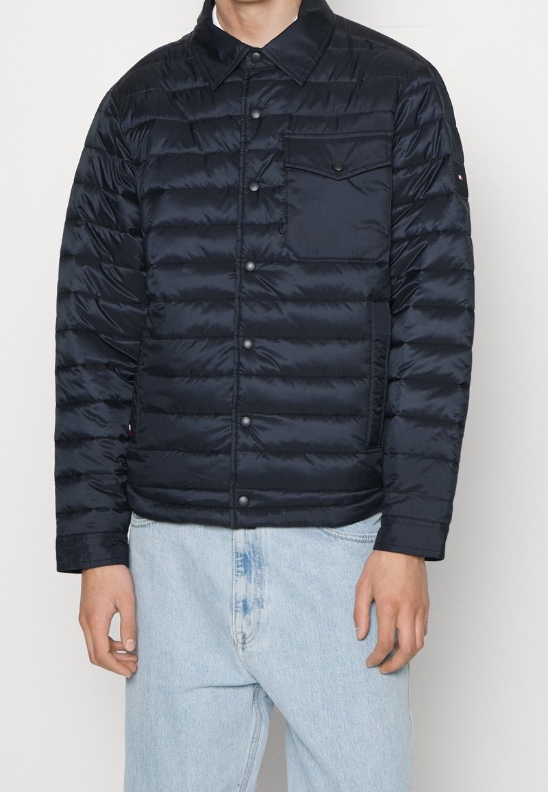 Mężczyzna w ciemnoniebieskiej pikowanej kurtce zapinanej na guziki z kieszenią na piersi oraz jasnoniebieskich dżinsach, stojący na jednolitym tle.