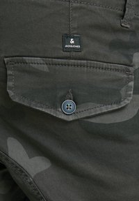 Kamuflážová kapsa na kalhotách z tmavě zelené látky s černou etiketou „JACKS & JONES“, zapnutá modrým knoflíkem a viditelným šitím.