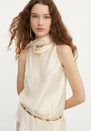 Jonge vrouw met lang krullend haar, die een mouwloos crèmekleurig topje draagt, een gouden en amberkleurige kettingriem en een enkele lange gouden oorbel.