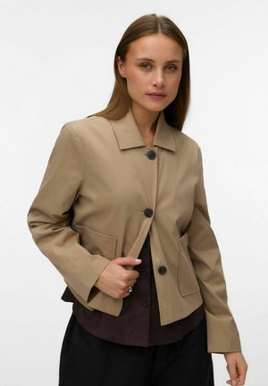 Femme portant une veste beige boutonnée sur un haut sombre, cheveux longs et raides, regardant la caméra sur un fond blanc uni.