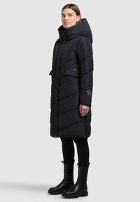 Must soe-parka kõrge kraega, siksakmustriga, esiteljekotid ja küljetõmblukud. Kantud mustade retuuside ja pahkluu pikkuste saapaididega.