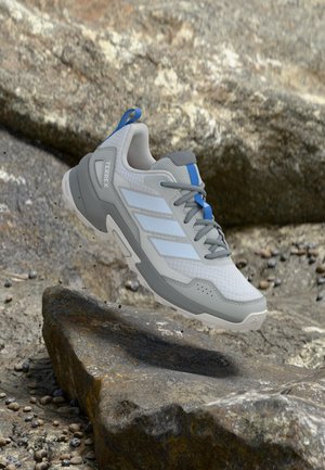 Sivo-plava planinarska cipela Adidas Terrex visi u zraku iznad kamenitog terena s malim rasutim kamenčićima.