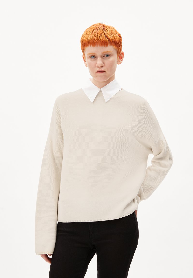 Beige Strickpullover mit lockerer Passform und langen Ärmeln, unter dem ein weißes Hemd mit Kragen getragen wird. Kombiniert mit schwarzen Hosen, vor einem weißen Hintergrund.