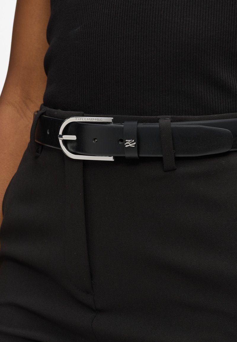 Cinturón de cuero negro con una hebilla de metal plateado, que presenta un lazo y un detalle grabado. El cinturón se lleva con ropa negra.