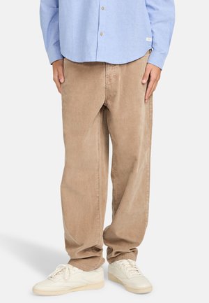 Beige corduroy pants with relaxed fit, featuring vertical ribbing. Paired with white sneakers and a light blue button-up shirt.