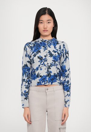 Kvinne iført en grå knappet cardigan med blå blomsterdesign og lyse beige bukser med lommer og glidelåsdetaljer.