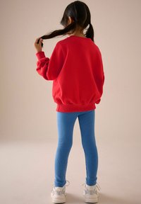 Rød oversized sweatshirt med samlet kant, matchet med blå ribbede leggings og hvide sneakers, set fra bagenden. Langt mørkt hår i hestehaler.
